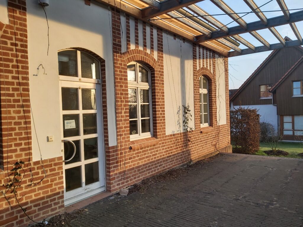 Das Bild zeigt einen Eingang zu einem Gebäude mit einer Backsteinfassade. Der Eingang besteht aus einer weißen Tür mit mehreren Fensterscheiben, die von zwei ebenfalls weißen Fenstern flankiert wird. Über der Tür und den Fenstern sind weiße Putzflächen, die von vertikalen roten Backsteinstrukturen eingerahmt sind. Ein Teil der Fassade ist von Kletterpflanzen bedeckt. Im Hintergrund sind weitere Gebäude zu sehen, die teilweise aus Holz gefertigt sind, sowie Büsche und ein gepflegter Rasen. Der gesamte Bereich ist von der Abendsonne beleuchtet, was eine warme und einladende Atmosphäre schafft.