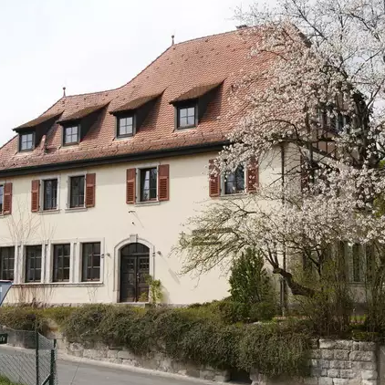 Das Bild zeigt ein großes weißes Gebäude mit Sprossenfenstern und Fensterläden. Auf dem Dach befinden sich kleine Gauben. Vor dem Haus sind Bäume, Sträucher und andere Grünpflanzen zu sehen.