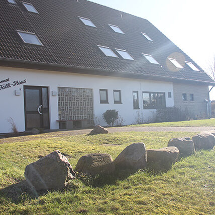 Das Bild zeigt ein großes weißes Haus . Vor dem Haus befindet sich eine Wiese mit großen Steinen.