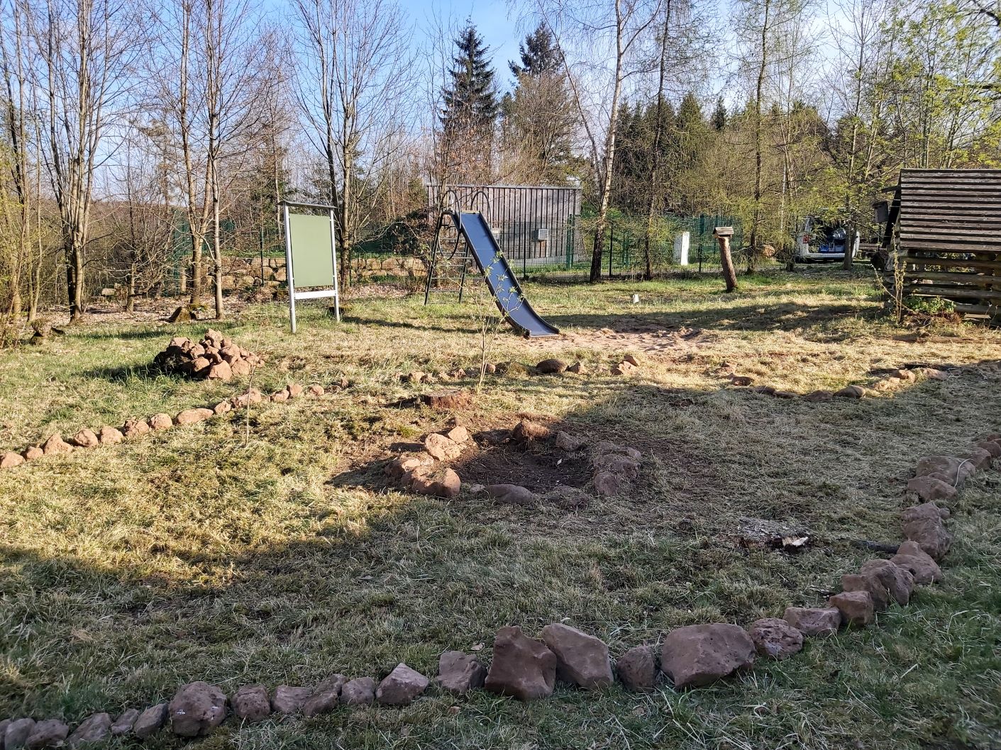 Auf dem Foto befindet sich ein großes Außengelände. Es ist mit Gras bewachsen und mit Steinen begrenzt. Eine Feuerstelle, eine Rutsche, eine Tafel und mehrere Bäume sind zu sehen.