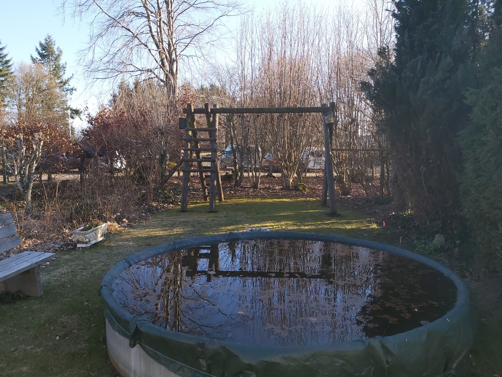 Im Vordergrund des Bildes ist ein abgedeckter Pool auf einer Wiese zu sehen. Im Hintergrund befindet sich eine ältere Holzschaukel und Bäume.