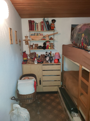 Auf dem Foto ist in kleiner Raum mit einem Stockbett zu sehen. An der weißen Wand steht eine helle Kommode mit unzähligen alten Stofftieren, darüber ein Regal mit alten Büchern und Spielsachen.