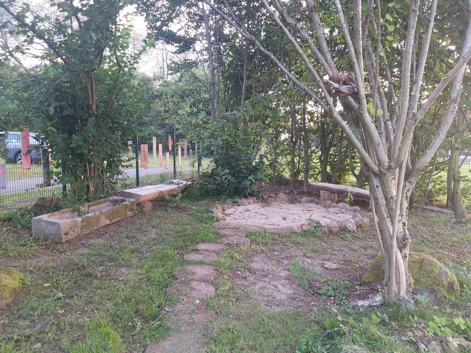 Auf dem Bild ist ein Grundstück mit vielen Bäumen und Sträuchern zu sehen. In der Mitte befindet sich ein Sandplatz, der mit Natursteinen begrenzt ist. Links und rechts davon stehen Holzbänke. Zu dem Sandplatz führt ein Weg aus Baumscheiben. Im Hintergrund ist ein Zaun mit bunt bemalten Brettern zu sehen.