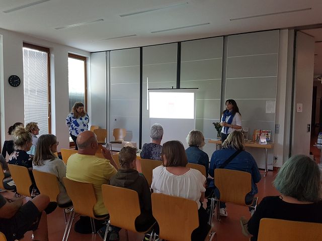 Eine Gruppe von Menschen sitzt in einem Raum auf Stühlen und hört einer Präsentation zu. Eine Frau steht am Projektorbildschirm, während die andere links von ihr steht und spricht. Der Raum hat große Fenster auf der linken Seite und eine Uhr an der Wand.