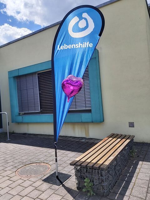 Das Bild zeigt eine blaue Fahne mit der Aufschrift "Lebenshilfe". Die Fahne steht vor einem Gebäude mit hellgelber Wand und türkisfarbenen Fensterrahmen. Neben der Fahne befindet sich eine Parkbank. An der Fahne ist ein pinker Herzballon befestigt.