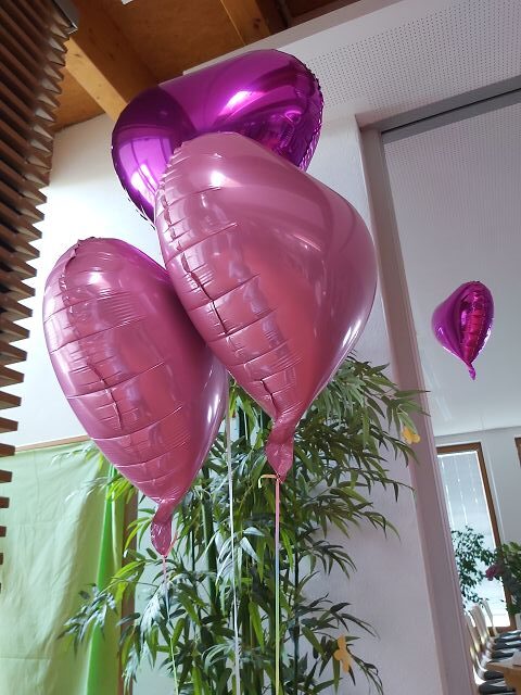 Das Bild zeigt drei rosa herzförmige Luftballons, die in einem Innenraum schweben. Im Hintergrund sind Pflanzen und ein Fenster zu sehen.