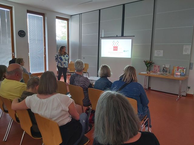 Eine Frau hält einen Vortrag vor einer Gruppe von Menschen in einem Konferenzraum. Eine Projektionsleinwand zeigt eine Präsentation. Die Zuhörer sitzen auf Stühlen und hören aufmerksam zu. Der Raum ist hell und modern eingerichtet.