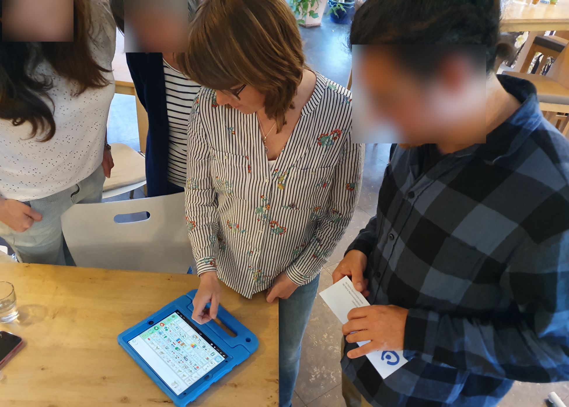 Mehrere Personen stehen um einen Tisch. Auf dem Tisch liegt ein Tablet mit einer blauen Schutzhülle, auf dessen Bildschirm verschiedene Symbole zu sehen sind. Eine Person bedient das Tablet, während eine andere Person ein Blatt Papier hält. Im Hintergrund sind weitere Tische und Stühle zu erkennen.