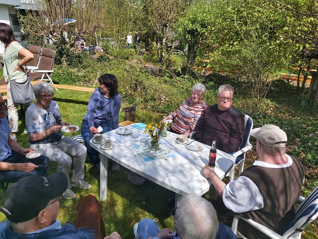 Eine Gruppe von Menschen sitzt um einen Tisch im Freien. Es gibt Kuchen, Tassen und Teller auf dem Tisch sowie einen Blumenstrauß in der Mitte. Die Personen scheinen sich zu unterhalten und eine angenehme Zeit im Garten zu verbringen. Im Hintergrund ist grüne Vegetation und ein Teil eines Hauses zu sehen.