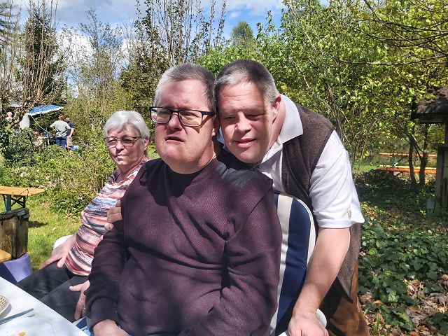 Eine Gruppe von Menschen sitzt im Freien an einem Tisch. Die Umgebung ist grün, sie befinden sich in einem Garten. Im Hintergrund sind andere Personen und Bäume zu sehen. Das Wetter ist sonnig und angenehm.