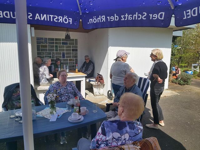 Eine Gruppe von Menschen sitzt und steht gemütlich im Freien unter einem großen blauen Sonnenschirm. Auf dem Tisch befinden sich Teller, Tassen und eine Flasche. Im Hintergrund sind mehrere Personen in einer Nische zu sehen. Die Atmosphäre wirkt entspannt und gesellig.