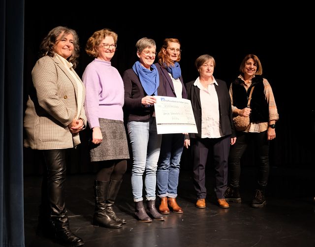 Eine Gruppe von sechs Frauen steht dicht nebeneinander auf einer Bühne. Zwei Personen in der Mitte halten einen großen Scheck in den Händen. Sie tragen überwiegend legere, aber stilvolle Kleidung. Alle lächeln freundlich.