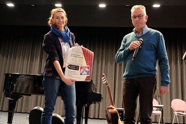 Zwei Personen stehen auf einer Bühne. Die Frau hält ein Plakat, während der Mann ein Mikrofon in der Hand hat. Im Hintergrund sind ein Klavier und eine Gitarre zu sehen.