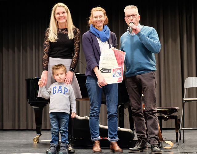 Vier Personen stehen auf einer Bühne. Eine Frau hält die Hand eines kleinen Jungen. In der Mitte hält eine zweite Frau ein großes Papier oder Plakat in den Händen. Ein Mann steht daneben und zeigt einen Daumen nach oben. Hinter ihnen ist ein Flügel sichtbar.