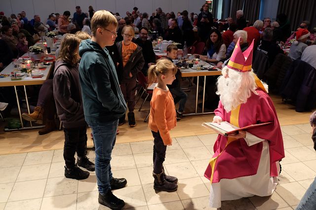 In einem festlich beleuchteten Raum stehen mehrere Kinder in einer Warteschlange vor einem als Nikolaus verkleideten Mann. Er trägt ein leuchtend rosa und goldenes Gewand und kniet sich zu einem kleinen Mädchen. Um ihn herum sind mehrere Tische gedeckt, an denen Menschen gemütlich sitzen und sich unterhalten. Es herrscht eine warme und einladende Atmosphäre.