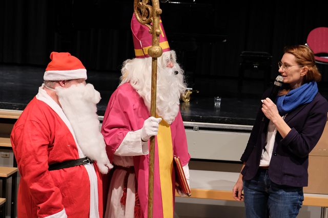 In dem Bild sind zwei Personen in festlicher Kleidung zu sehen. Eine Person ist als Weihnachtsmann verkleidet mit einem roten Mantel und weißem Bart. Die andere Person trägt ein bischöfliches Gewand mit einem langen, rosa Mantel und hält einen goldenen Bischofsstab. Beide stehen vor einer Bühne mit dunklem Hintergrund und unterhalten sich mit einer dritten Person, die ein Mikrofon hält und einen blauen Schal trägt.