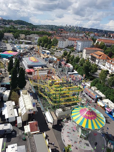 Das Bild zeigt ein Schützenfest aus der Vogelperspektive. Man sieht Fahrgeschäfte, Buden und Besucher verteilt auf dem Gelände. Im Hintergrund erstreckt sich eine städtische Landschaft mit Wohnhäusern und grünen Bäumen. Der Himmel ist leicht bewölkt.