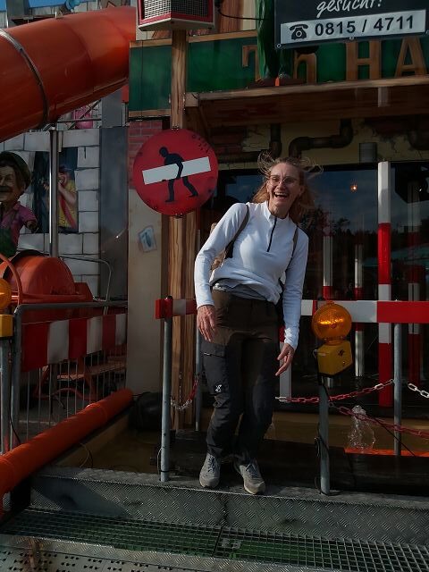 Ein lachende Frau steht vor einem Eingang eines Gebäudes auf einem Schützenfest. Das Gebäude hat eine bunte, verspielte Fassade mit verschiedenen Schildern und Dekorationen, darunter einer großen Rutsche. Die Frau trägt Outdoor-Kleidung, ihre Haare wehen im Wind und sie lehnt sich leicht nach rechts. Im Hintergrund sind reflektierende Oberflächen und bunte Lichter zu sehen.