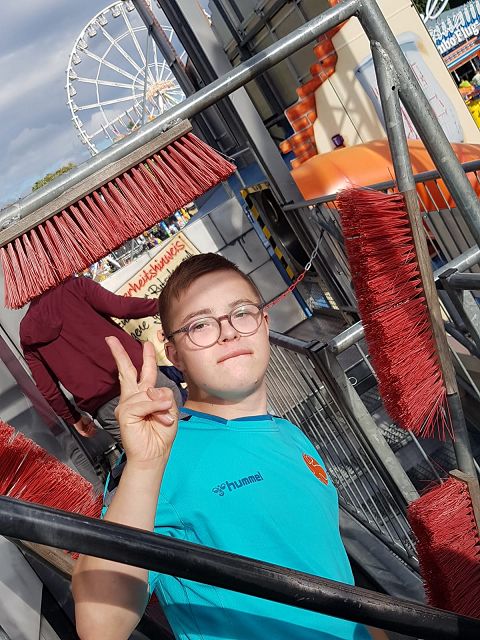 Ein junger Mann zeigt ein Victory- oder Peace-Zeichen. Im Hintergrund befindet sich ein großes Schützenfest mit einem Riesenrad und einer Rutsche. Insgesamt herrscht dort eine lebhafte und fröhliche Atmosphäre.