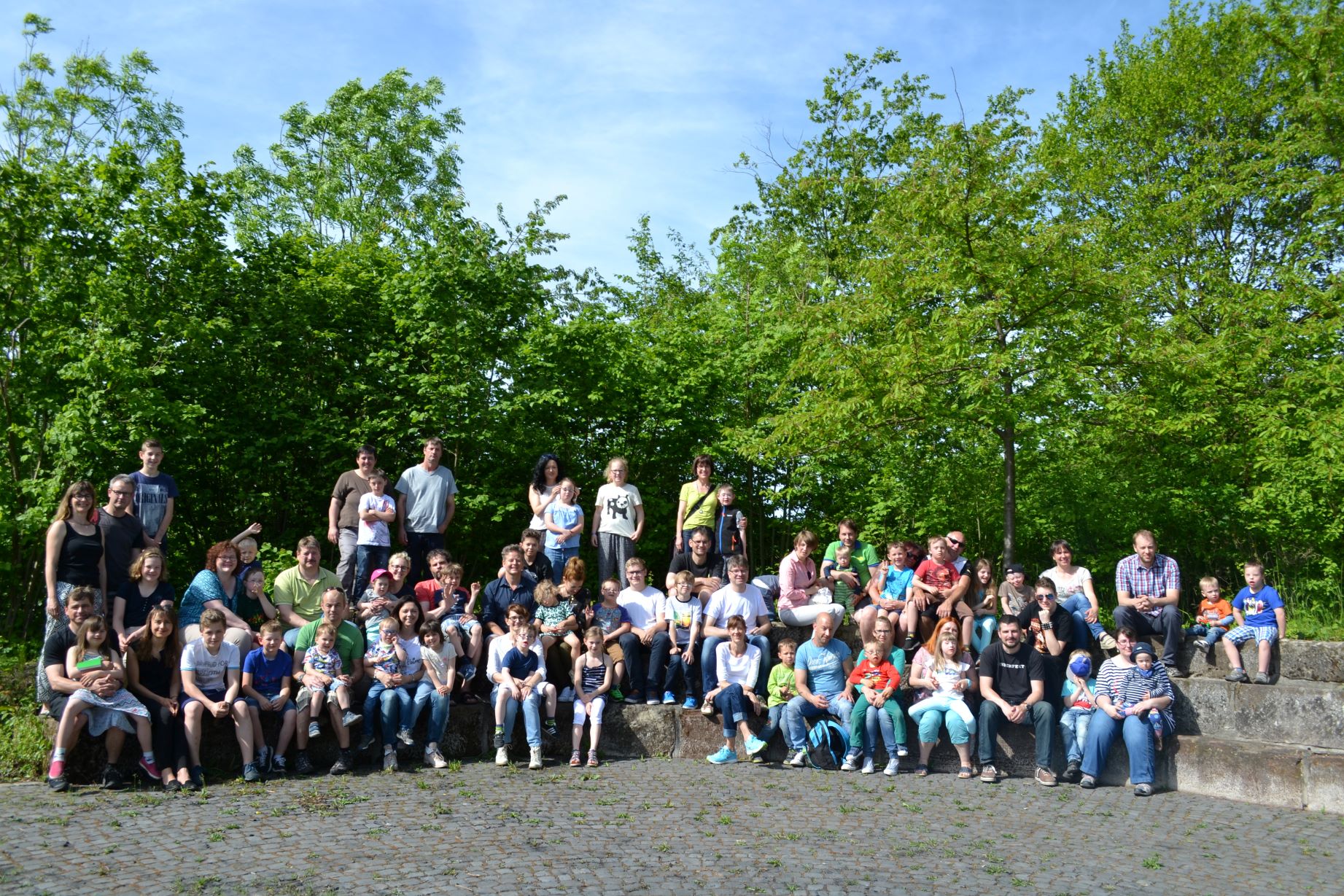 Auf dem Foto sind viele Familien zu sehen. Sie sitzen im Halbkreis auf großen Stufen im Hintergrund sieht man Bäume und blauen Himmel.