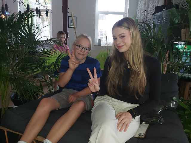 Das Bild zeigt eine junge blonde Frau und einen Jugendlichen mit Brille auf einem modernen Sofa. Sie zeigen ein Victory- oder Peace-Zeichen. Im Hintergrund sieht man einen weiteren Jungen mit Brille und einige Grünpflanzen.