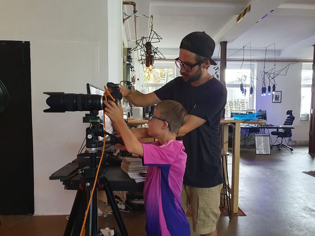 Ein junger Mann mit Kappe, Brille und Bart zeigt einem Jungen mit Brille und pinkfarbenen Trikot eine Fotokamera. Sie steht auf einem Stativ. Im Hintergrund sieht man eine moderne Büroeinrichtung.