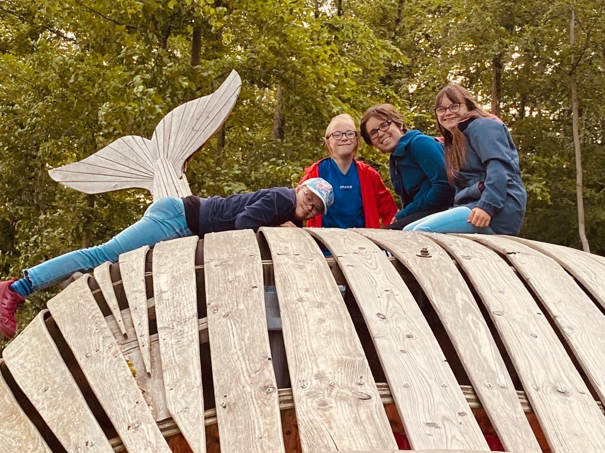 3 junge Menschen sitzen auf einem riesigen Wal aus Holz. Ein Mädchen mit Kappe liegt auf dem Holz. Alle 4 tragen eine Brille und lächeln fröhlich. Im Hintergrund sieht man Bäume und die Schwanzflosse des Wals aus Holz.