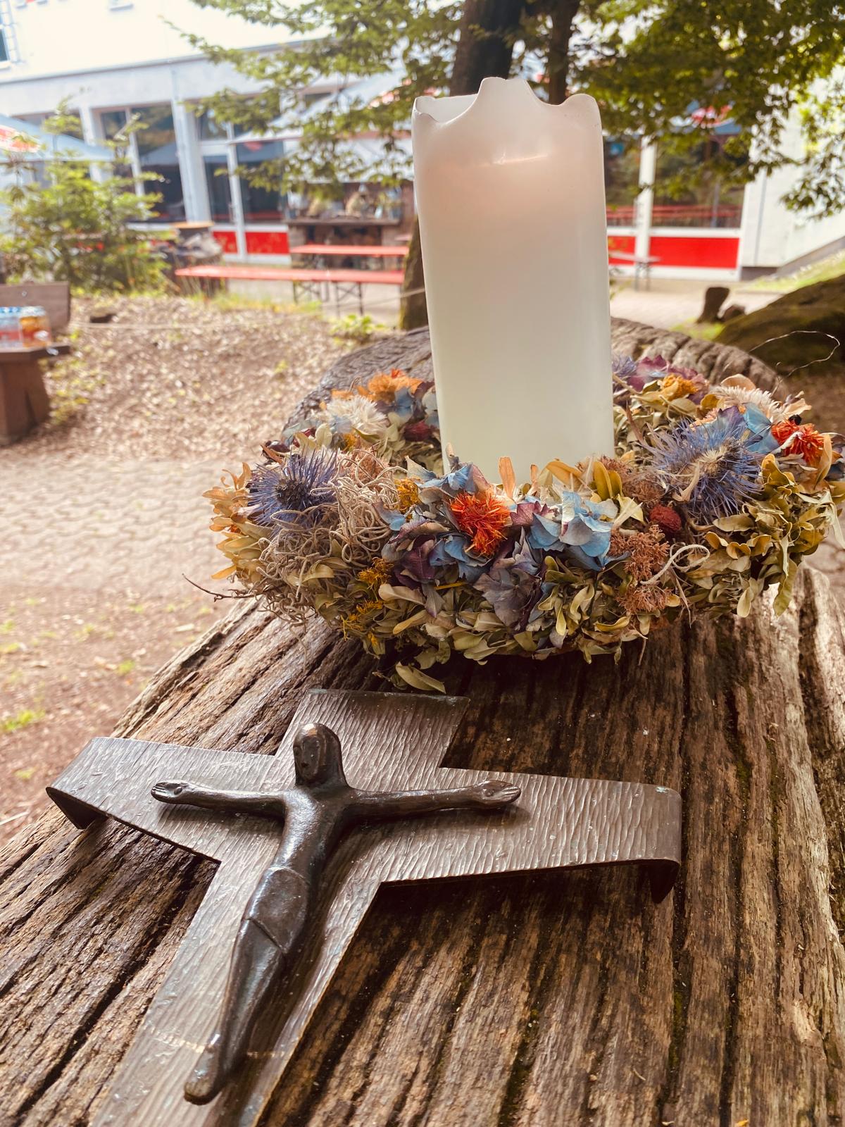 Auf einem dicken Baumstamm steht eine weiße brennende Kerze. Sie ist mit einem dicken Blumenkranz geschmückt. Davor liegt ein christliches Kreuz aus Metall. Im Hintergrund ist ein Gebäude zu sehen.