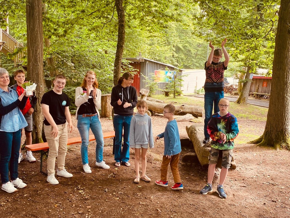 Auf dem Bild sind Kinder, Jugendliche und Erwachsene zu sehen. Sie klatschen und scheinen sich zu freuen. Im Hintergrund befinden sich viele Bäume.