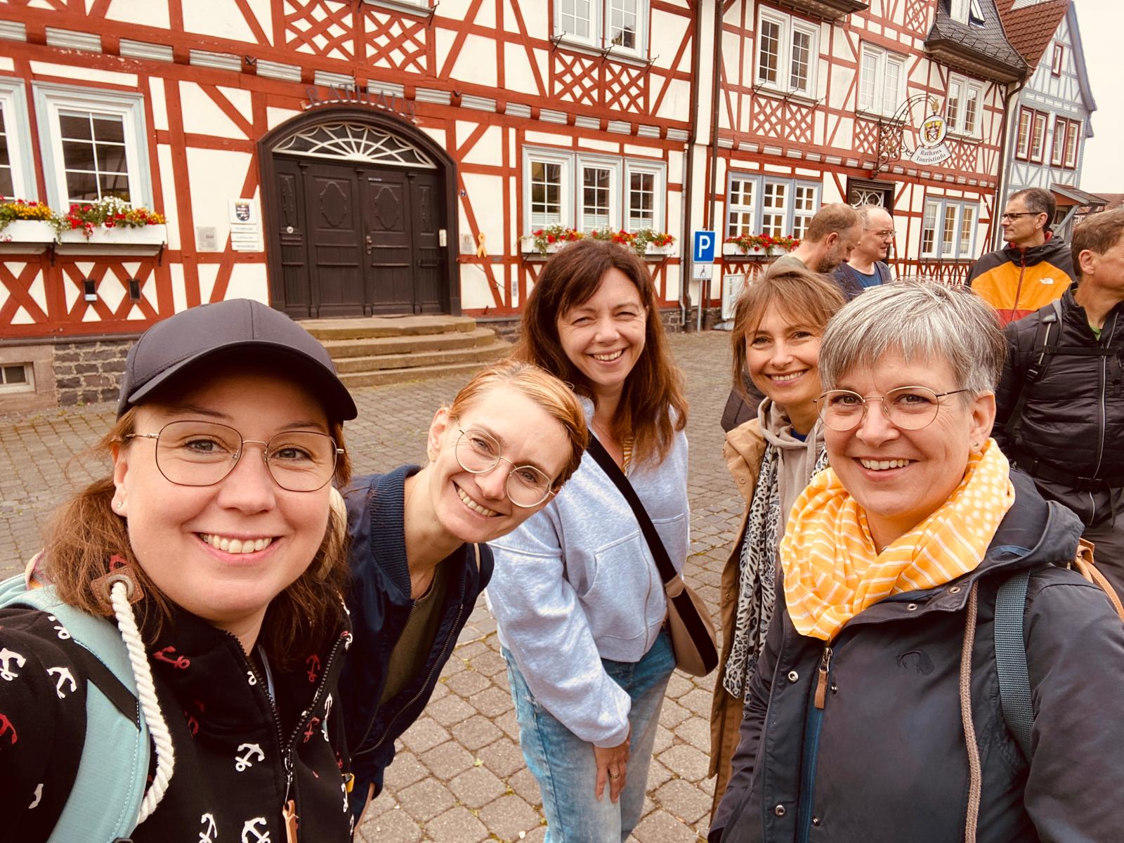 5 Frauen schauen lachend in die Kamera. Dahinter stehen ein paar Männer, die sich unterhalten. Im Hintergrund befindet sich ein großes Fachwerkhaus.