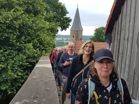 Auf dem Bild sind mehrere gut gelaunte Erwachsene zu sehen. Sie laufen entlang einer Mauer in einer Reihe hintereinander. Im Hintergrund ist ein historischer Turm mit spitzem Dach zu sehen.