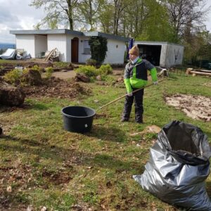 Ein Jugendlicher arbeitet im Garten und hält eine Gartenharke. Im Vordergrund stehen ein schwarzer Eimer und ein großer Müllsack auf einer Grasfläche. Im Hintergrund sieht man ein weißes Gebäude, Bäume und Geräte. Es scheint ein typischer Frühlingstag zu sein.