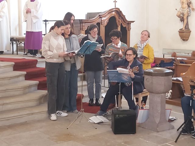 Das Foto zeigt eine Gruppe von Menschen, die in einer Kirche singen. Eine Frau spielt Gitarre, die anderen singen aus Notenblättern.