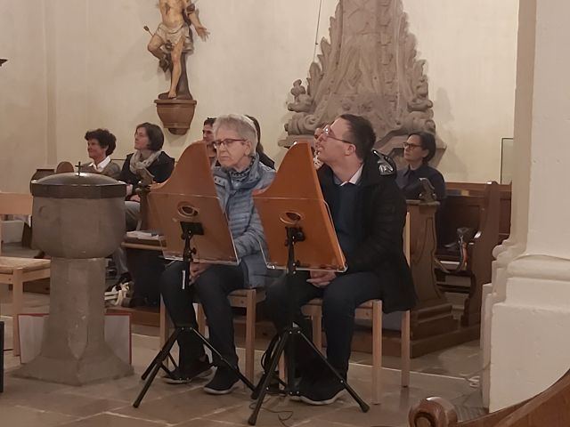 Das Bild zeigt eine Frau und einen Mann an einer Veh-Harfe. Im Hintergrund sitzen weitere Personen. Sie befinden sich in einer Kirche.