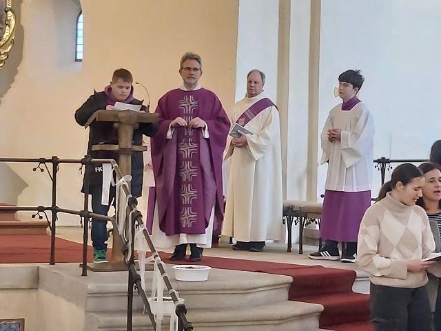 Das Bild zeigt eine Gruppe von Menschen, die in einer Kirche stehen. Es gibt einen Priester, der eine lilafarbene Robe trägt, einen anderen Mann in einer weißen Robe und zwei weitere Männer, die ebenfalls lilafarbene Roben tragen. Im Vordergrund steht ein junger Mann, der ein Mikrofon hält.