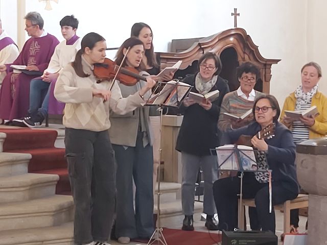 Das Bild zeigt eine Gruppe von Menschen in einer Kirche, die singen und Musik machen. Eine Frau spielt Geige, eine andere Gitarre und die restlichen Personen singen aus Gesangbüchern. Im Hintergrund sitzt ein Priester in einem violetten liturgischen Gewand.