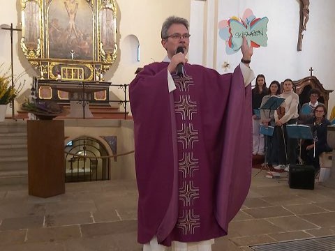 Das Bild zeigt einen Priester in einem violetten Gewand, der in einer Kirche steht und in ein Mikrofon spricht. Er hält ein Plakat mit der Aufschrift "Geben" in der Hand, das aussieht wie ein Schmetterling. Im Hintergrund sieht man einen Chor mit Notenständern, der auf den Priester blickt.