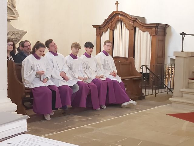 Das Bild zeigt sechs Jugendliche, die in einer Kirche auf einer Bank sitzen. Sie tragen weiße Gewänder mit violetten Untergewändern. Die Jugendlichen befinden sich in einer Kirche. Es scheinen Messdiener zu sein.