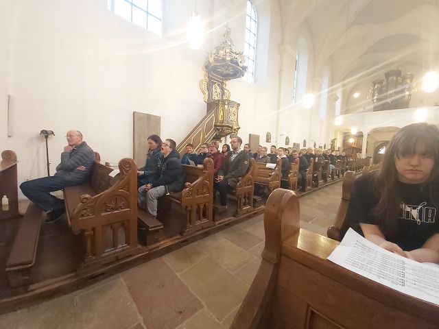 Es sind viele Menschen zu sehen. Sie sitzen in einer Kirche. Im Hintergrund ist eine Kanzel zu sehen.