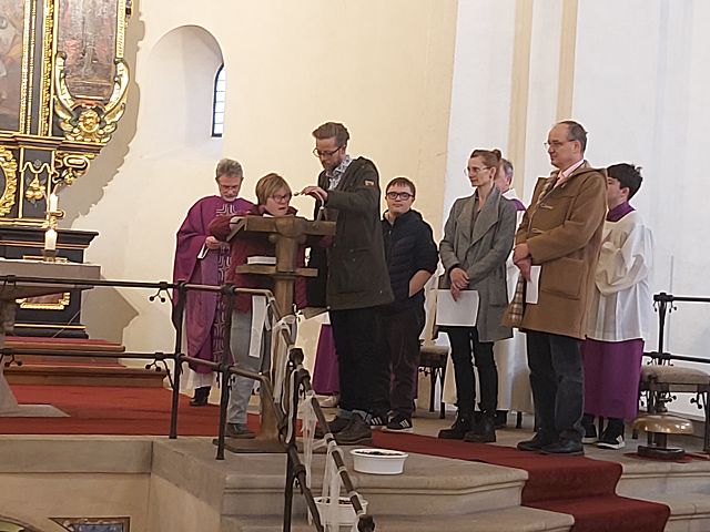 Das Bild zeigt mehrere Menschen in einer Kirche. Sie stehen um ein Ambo und halten Zettel in der Hand. Eine Frau steht am Mikrofon und liest etwas vor. Im Hintergrund sieht man einen Priester in violettem Gewand und Messdiener.