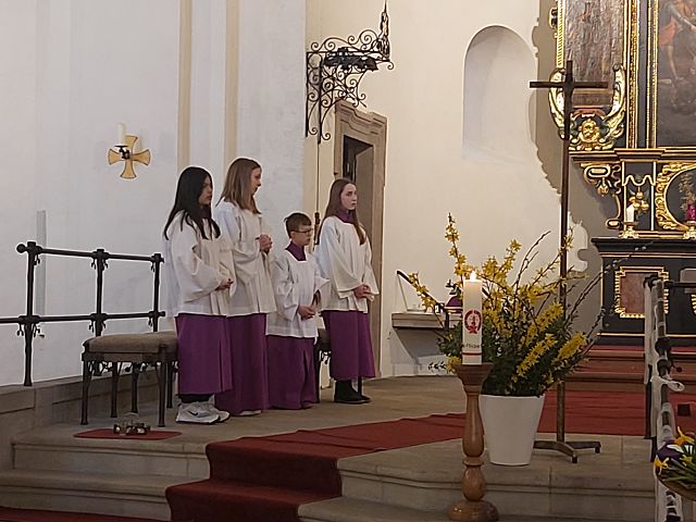 Das Bild zeigt 3 Mädchen und einen Jungen in einer Kirche. Sie tragen weiße Gewänder mit violetten Untergewändern. Es scheinen Messdiener zu sein.