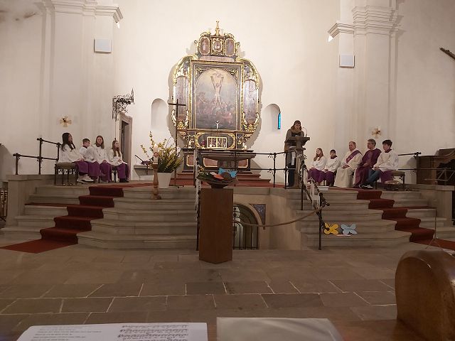 Es ist ein Altarraum zu sehen. In der Mitte befindet sich ein großes Gemälde mit goldenem Rahmen. Rechts der Mitte steht eine Frau am Ambo und spricht. Seitlich sitzen ein Priester und mehrere Messdiener in Gewändern. Im Vordergrund sieht man mehrere Stufen, die zu dem Altar hinauf führen.