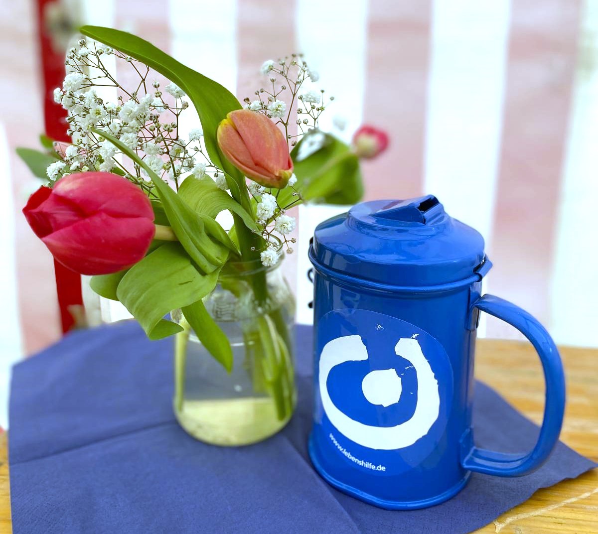 Auf dem Bild ist eine blaue Spendendose mit dem Logo der Lebenshilfe zu sehen. Daneben steht eine Blumenvase mit roten Tulpen. grünen Blättern und weißem Schleierkraut.