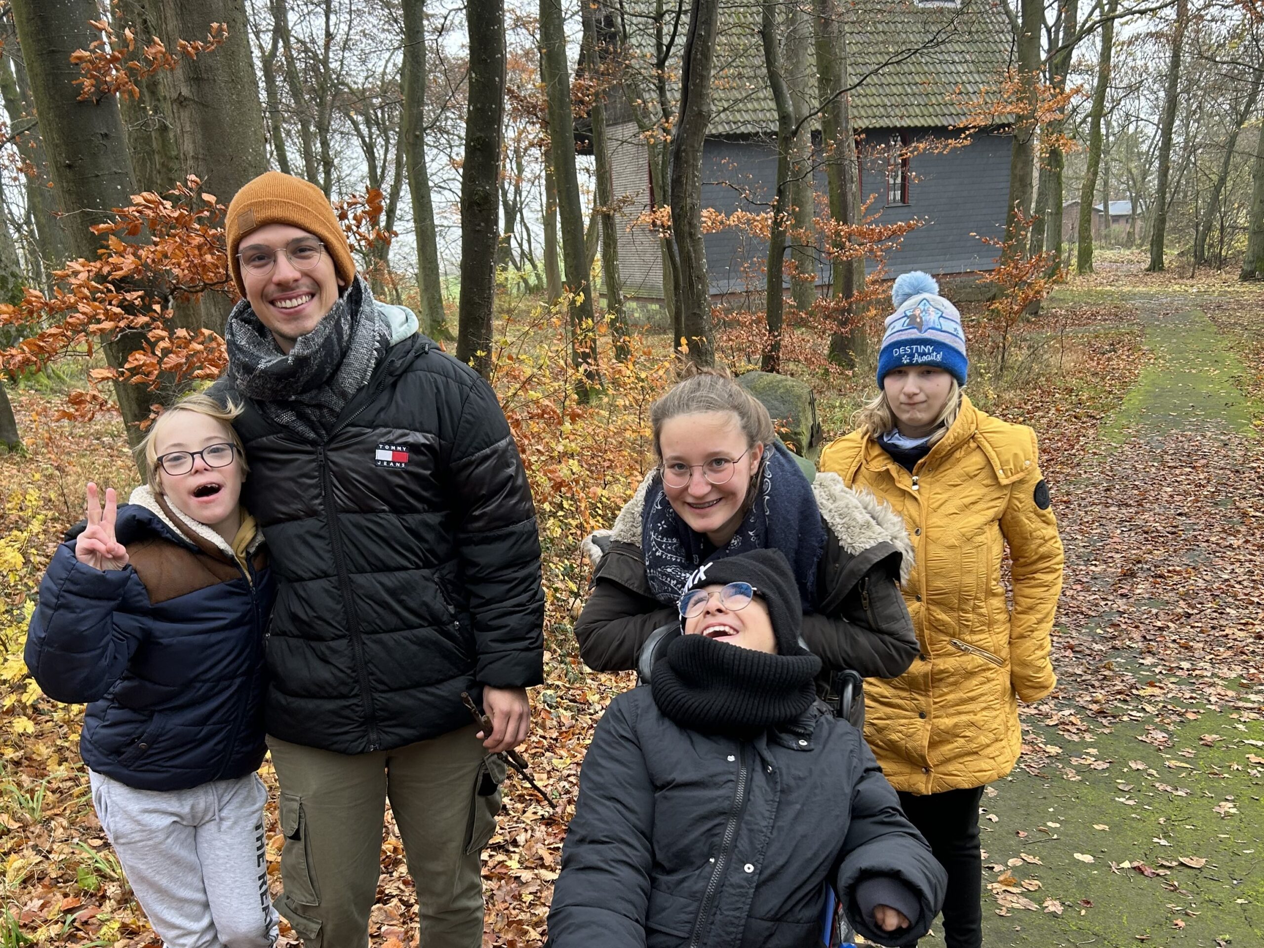 Auf dem Bild sind mehrere Jugendliche zu sehen. Im Vordergrund sitzt ein Junge im Rollstuhl, er hat eine rote Decke auf den Beine n liegen. Überall sind Bäume und Herbstlaub zu sehen. Alle lachen fröhlich.