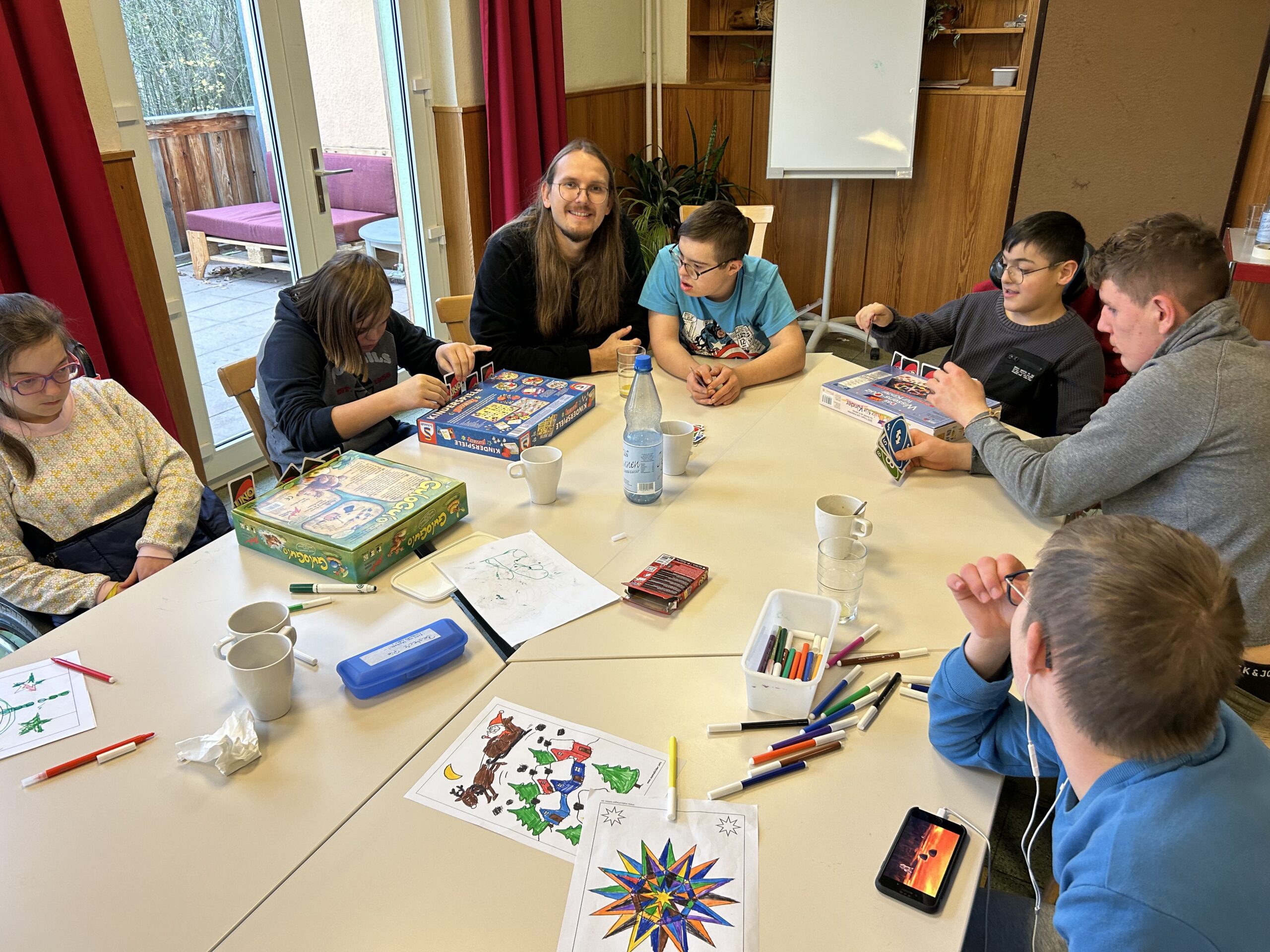 Auf dem Bild sind mehrere Jugendliche zu sehen. Sie sitzen an einem großen Tisch und spielen miteinander. Auf dem Tisch liegen verschiedene Spiele, Stifte und Bilder, außerdem Tassen und eine Wasserflasche.