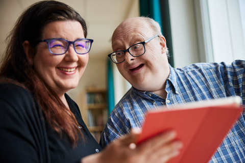 Eine Frau und ein Mann sind zu sehen. Beide tragen eine Brille und lachen freundlich. Die Frau hält eine rote Mappe in der Hand.