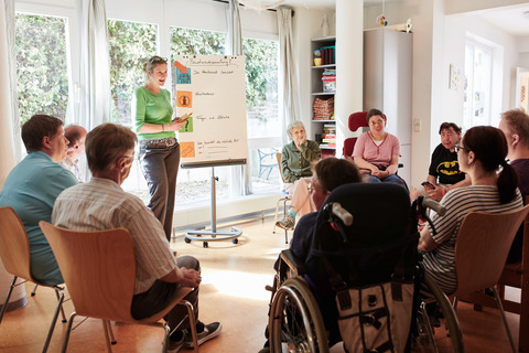 Auf dem Bild sind mehrere Erwachsene zu sehen. Sie sitzen im Kreis in einem hellen Raum. Am Rand des Kreises steht eine Frau neben einem Flipchart.