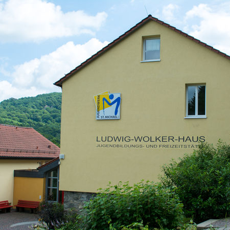 Es ist ein großes gelbes Haus mit einem Anbau zu sehen. Darauf steht in großer Schrift "Ludwig-Wolker-Haus" und darunter "Jugendbildungs- und Freizeitstätte". Links über dem Schriftzug befindet sich ein Plakat mit einem gelb-blauen Logo und dem Text Jugendwerk St. Michael.