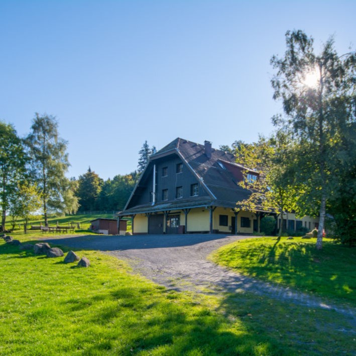 Auf dem Bild sieht man ein gelbes großes Haus mit einem 2-stöckigen Dach. Das Dach und der Giebel sind dunkelbraun. Ein Weg führt zu dem Haus. Rundherum sieht man Wiese und Bäume, die Sonne scheint.