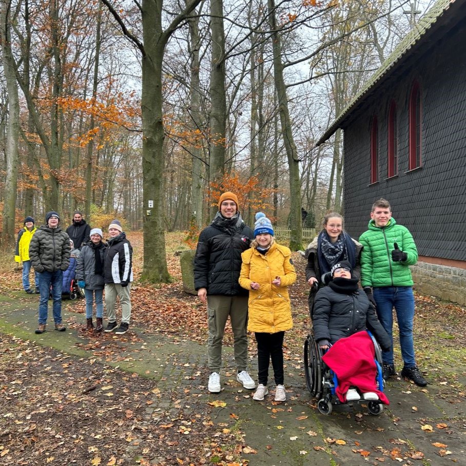 Das Bild zeigt mehrere Jugendliche. Sie sind mit dicken Jacken und Mützen bekleidet. Ein Kind sitzt im Rollstuhl und hat eine rote Decke auf den Beinen liegen. Die Gruppe befindet sich in einem Herbstwald, rechts sieht man einen Teil eines dunklen Gebäudes.